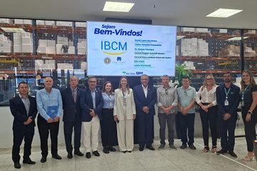 Grupo de doze pessoas posicionadas lado a lado posando para foto em frente a uma tela digital com mensagem de boas-vindas. Na tela constam os nomes de Daniel Lopes dos Santos, Clarice Terezinha Pasqualotto, Dr. Djeison Falavigna, Coronel Rogério Schmidt Navarro, Coronel Marco Antônio Moura dos Santos, Eder André e Letícia Wieczynski. Ao fundo, através de uma divisória de vidro, vê-se um galpão logístico com prateleiras cheias de caixas e suprimentos.